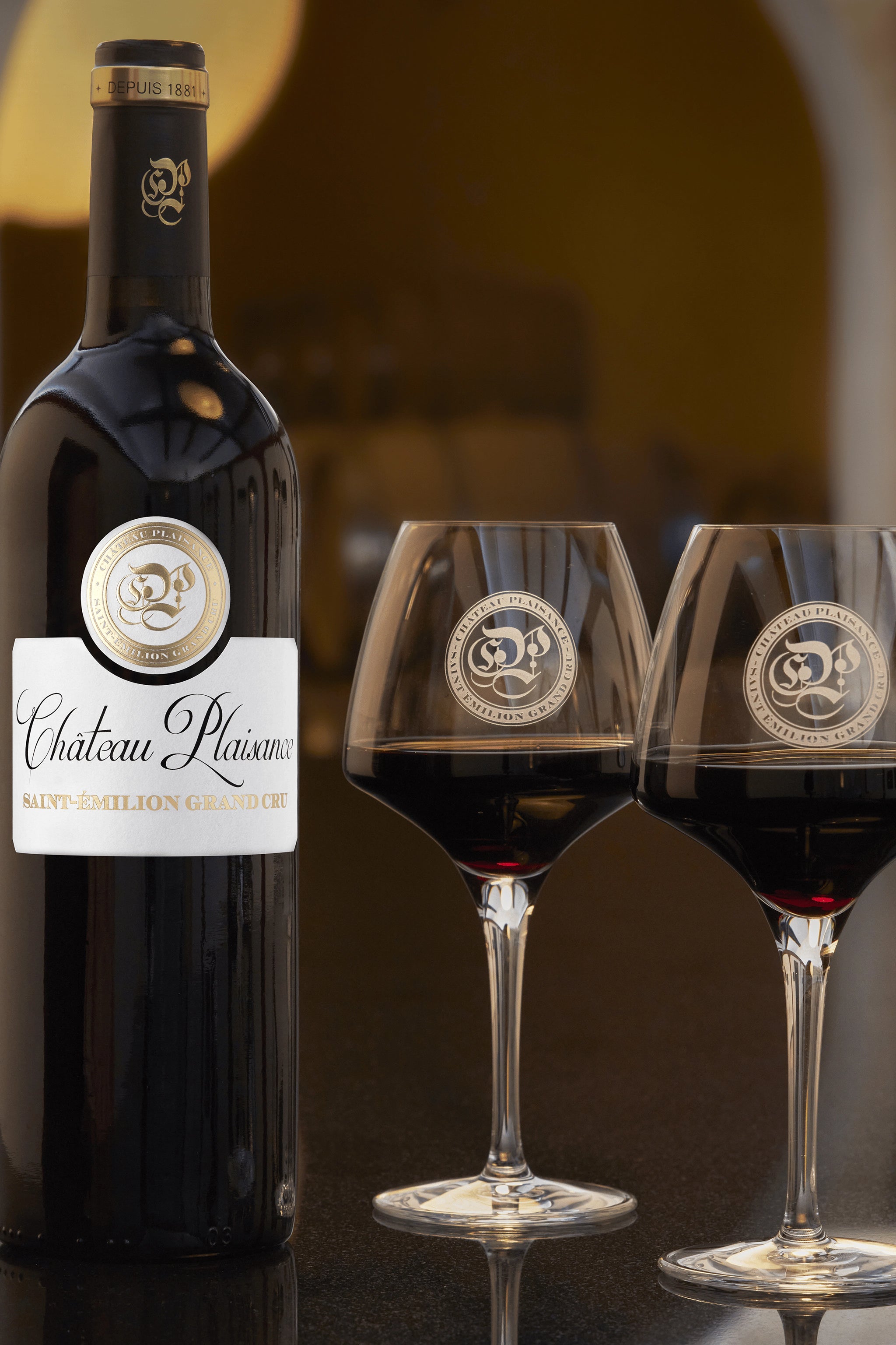 A photograph of a bottle of Château Plaisance award winning Saint-Émilion Grand Cru red wine with two glasses on the Château's tasting table.